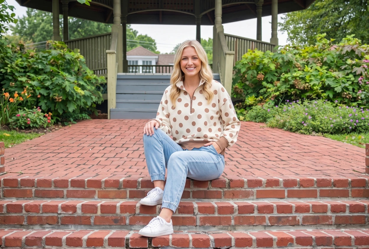 A woman sits on red brick steps in front of a wooden gazebo, wearing a simply southern cream pullover with brown polka dots, light blue jeans, and white sneakers.